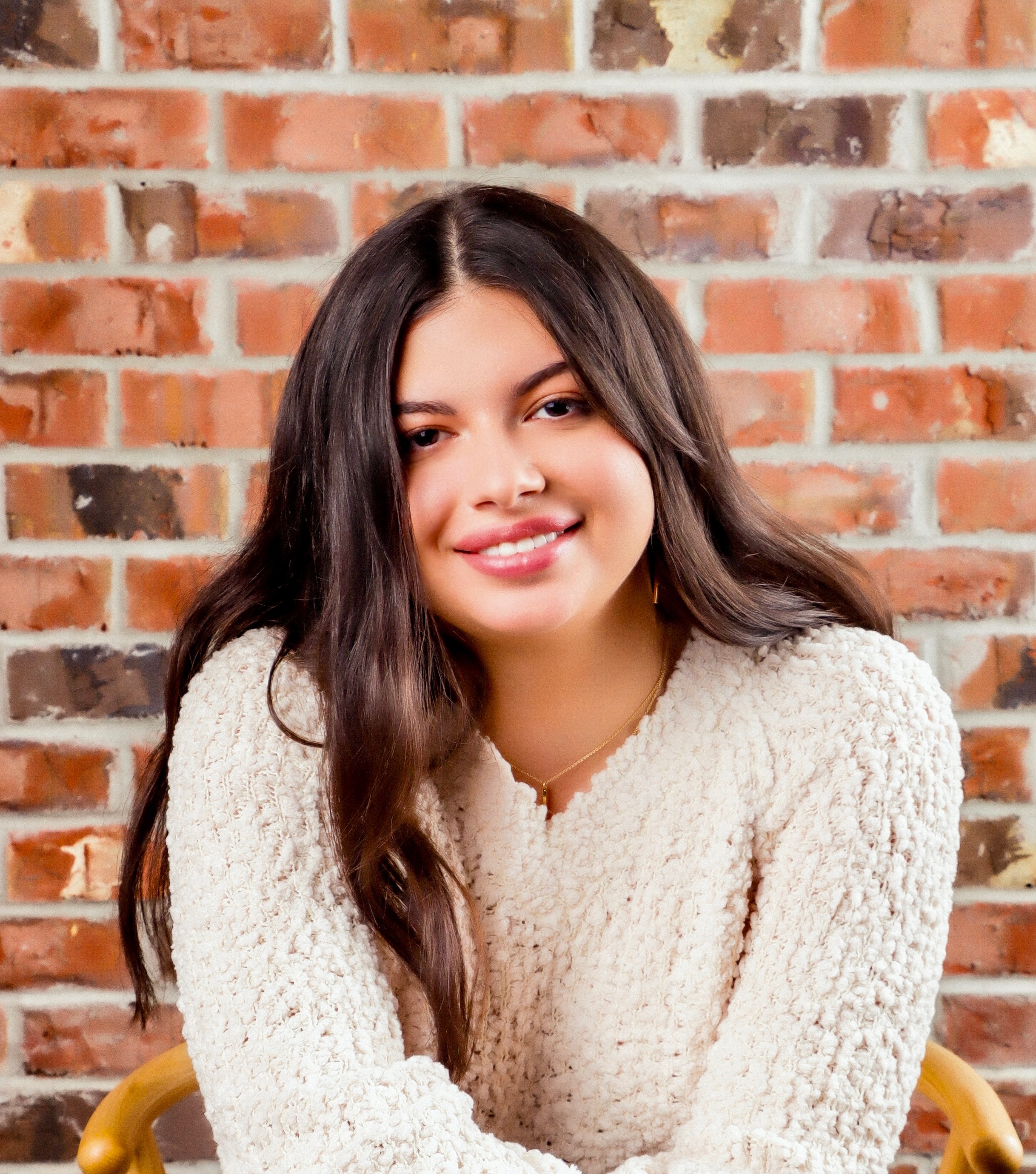 Photograph of Sabrina Califano smiling in front of a red brick wall. She has long dark hair and is wearing a cream-colored knit sweater.