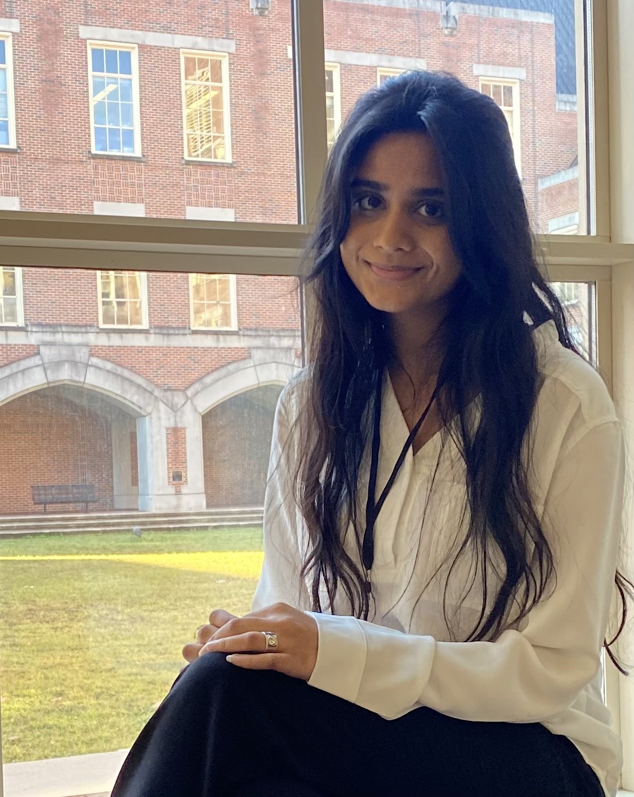 Photograph of Khushi Patel sitting by a large window with a brick building visible in the background. She has long dark hair and is wearing a white blouse.