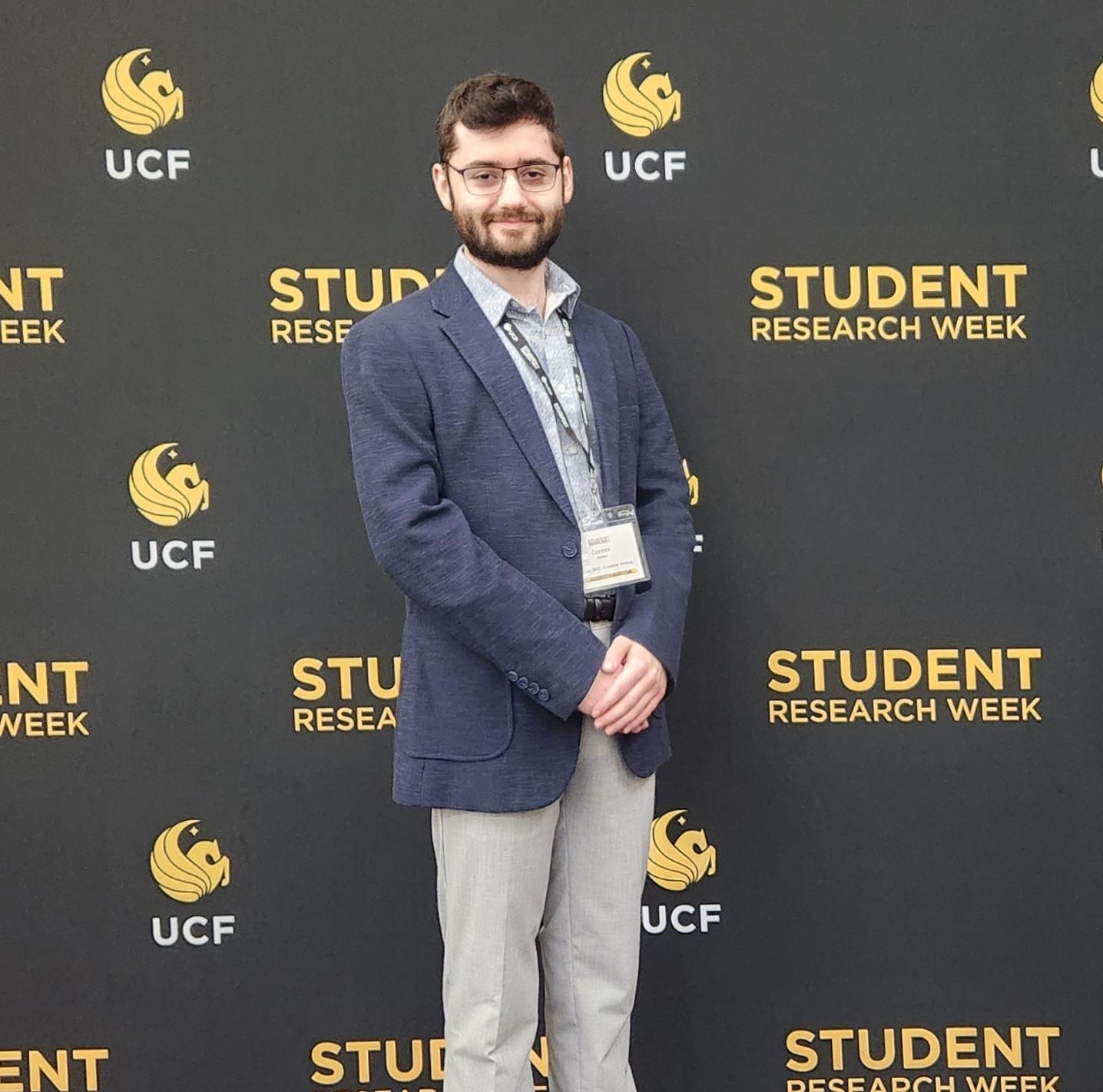 Photograph of Connor Seaton standing in front of a UCF Student Research Week backdrop. He is wearing a navy blazer and patterned button-down shirt, with glasses and a conference lanyard.